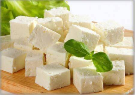 Cubes of Feta Greca on a wooden board with fresh basil and lettuce in the background.