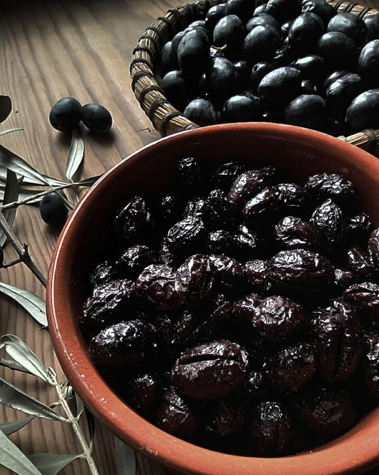 Olive Nere Secche Infornate e Condite Busta 3 kg in a bowl with fresh olives and olive leaves around.