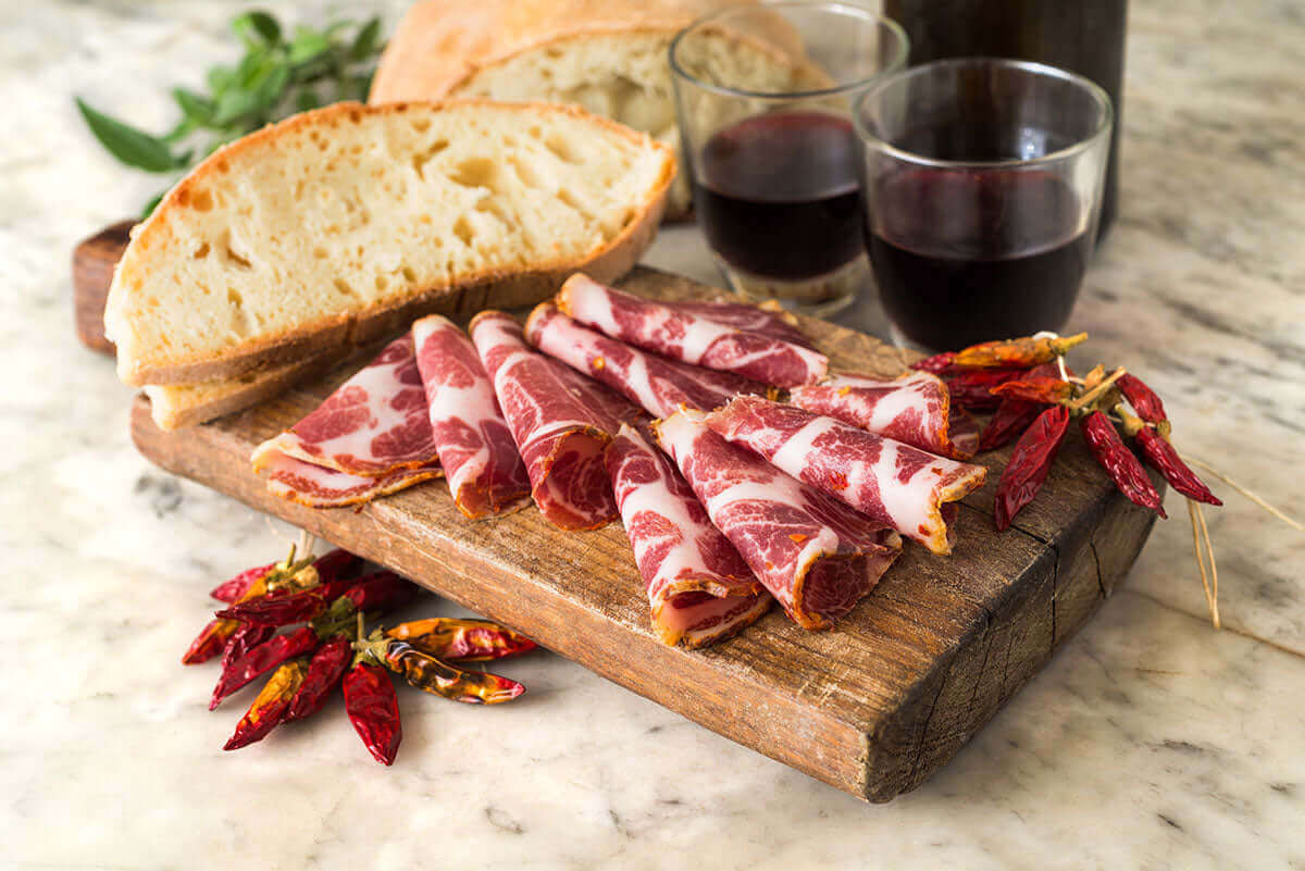 Capocollo - Artigianale di Calabria served on a wooden board with bread, wine, and red chili peppers.