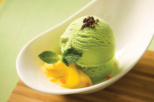 Green tea ice cream served with a sprig of mint and a dollop of mango sauce in a white dish on a wooden surface.
