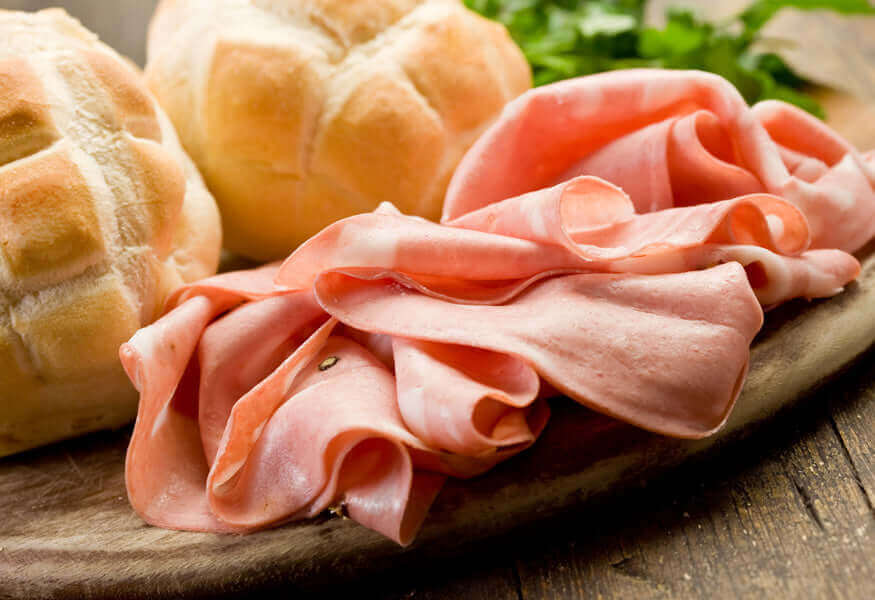 Mortadella al Cinghiale, thinly sliced meat on a wooden board with bread rolls and fresh herbs.