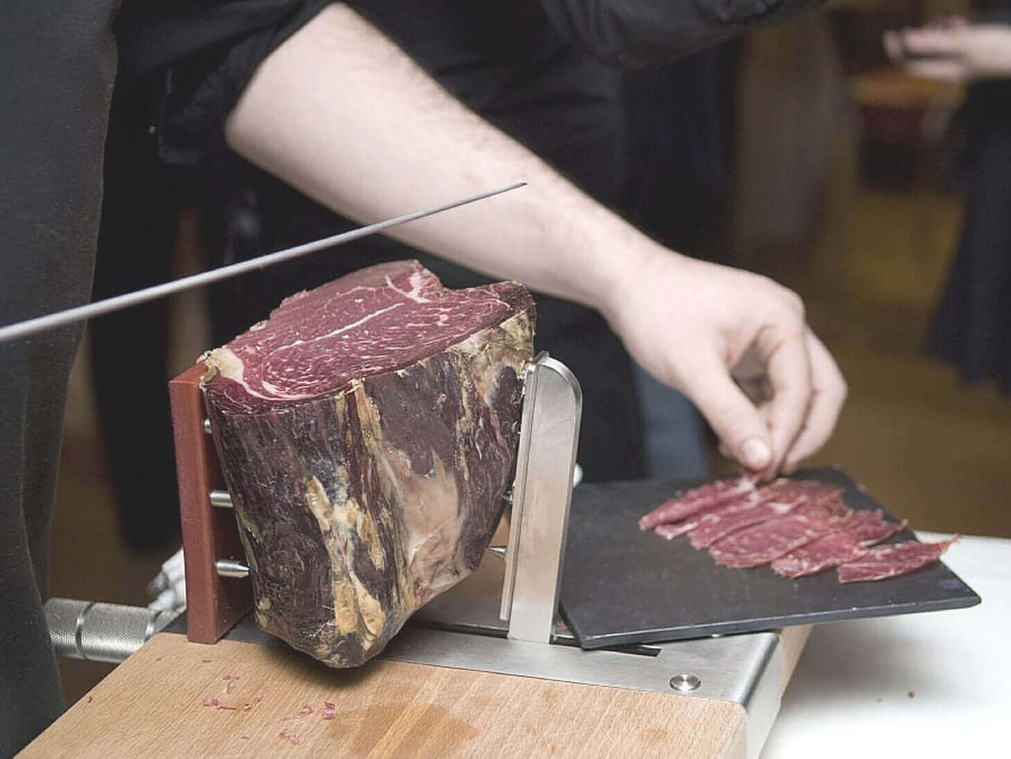 Person slicing cured meat with a manual slicer on a wooden table.