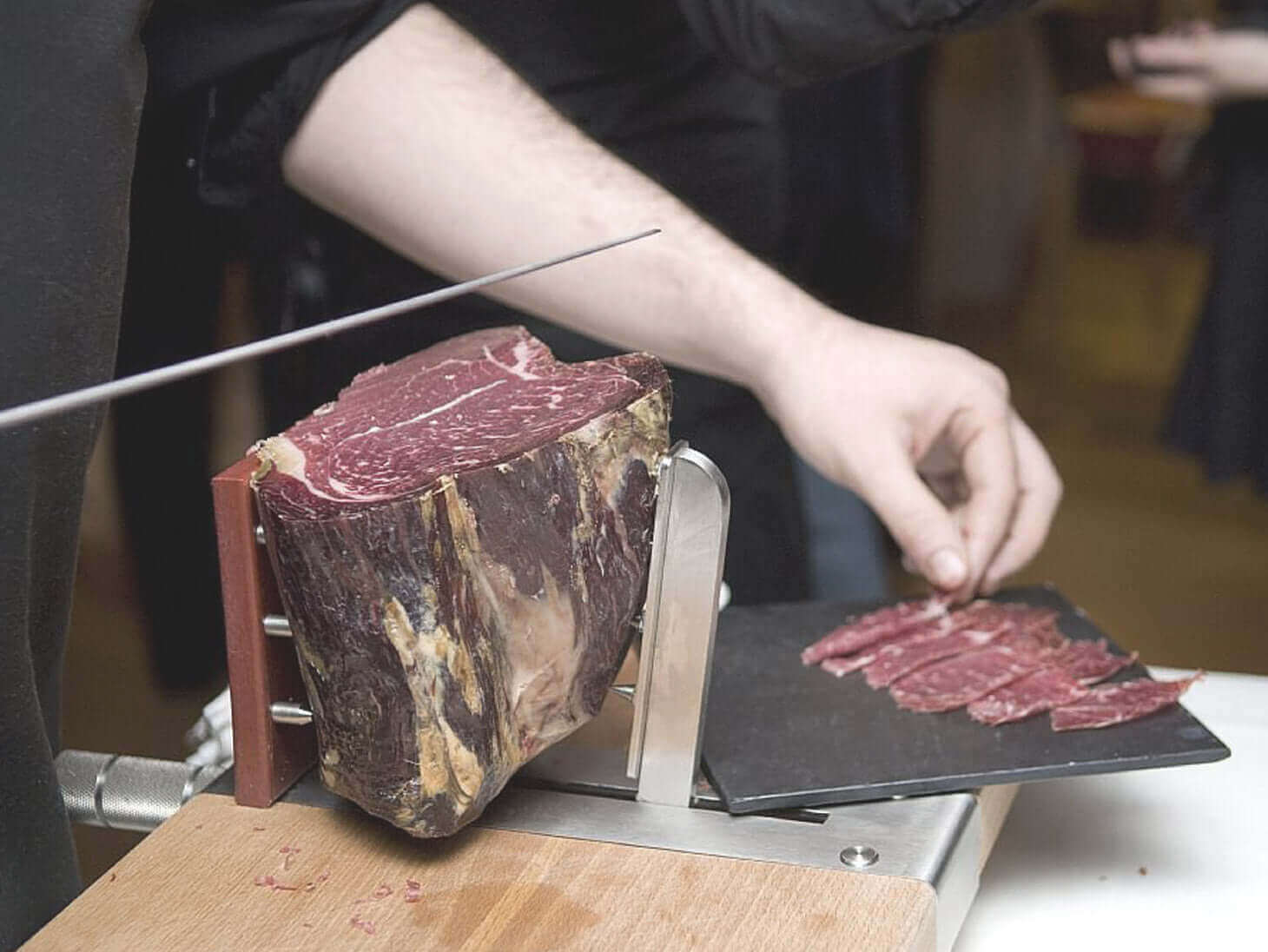 Person slicing cured meat with a manual slicer on a wooden table.